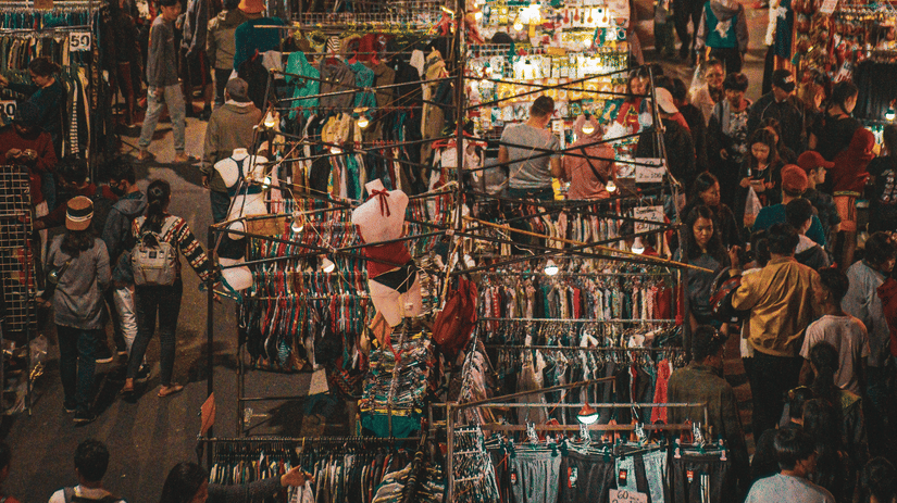 Bustling evening market filled with clothing stalls, shoppers, and vibrant shopping displays.