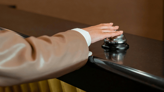 Person pressing a silver service bell on a wooden front desk counter.