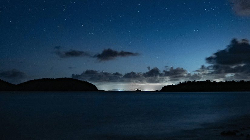 A sea beach with calm water under a starry sky with a few clouds