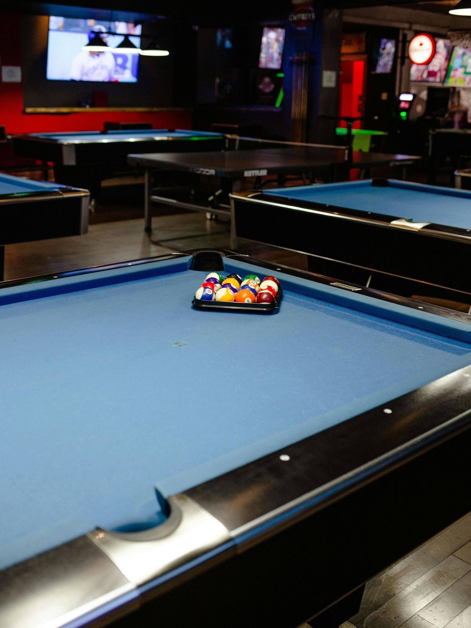 A dimly lit billiards room with several pool tables and a scoreboard in the background.