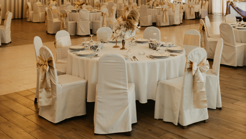A banquet hall set for a wedding reception with round tables in white linen, chairs dressed with gold sashes, crystal chandeliers, and floor curtains.