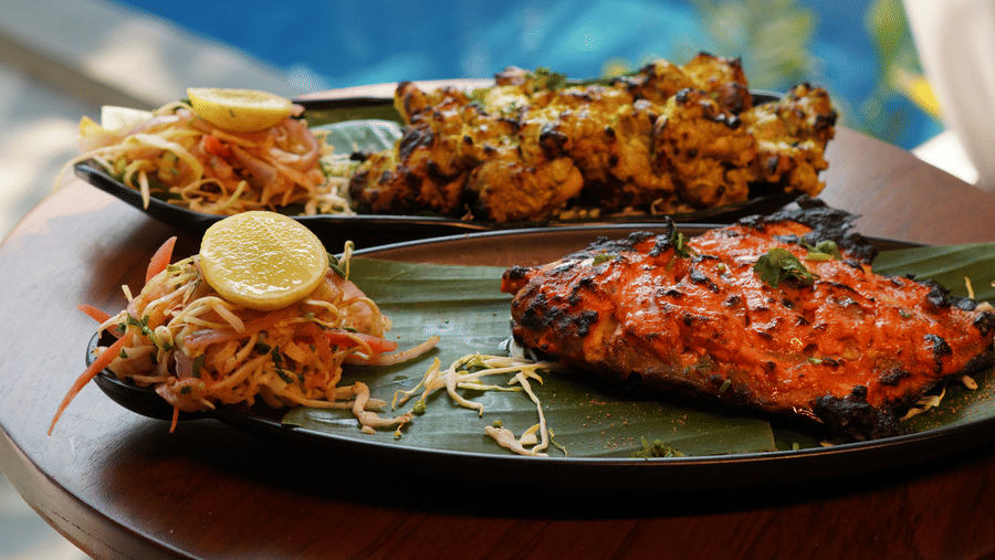 2 platters filled with grilled food, salads, and garnishes sit by a window overlooking a swimming pool at Bougainvillea, Alibaug.