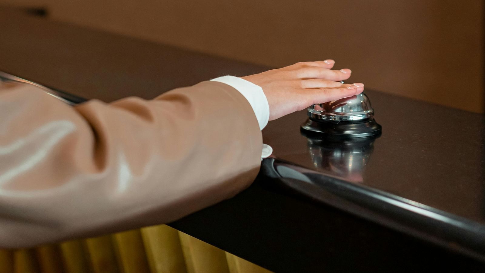 A hand in a light brown sleeve presses down on a silver reception bell on a dark counter.