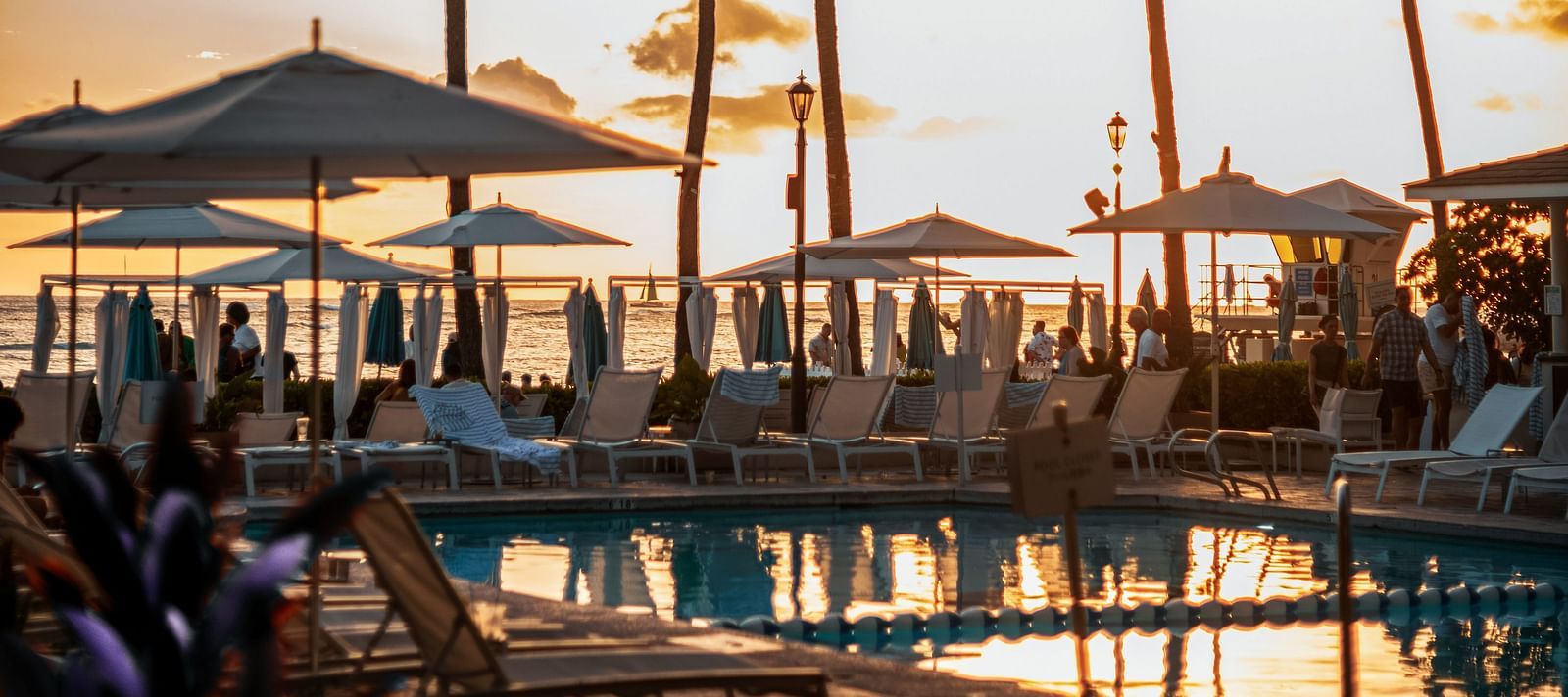 Lush tropical resort with a winding swimming pool, thatched-roof bungalows, and tall palm trees under a bright blue sky, suggesting a relaxing vacation.