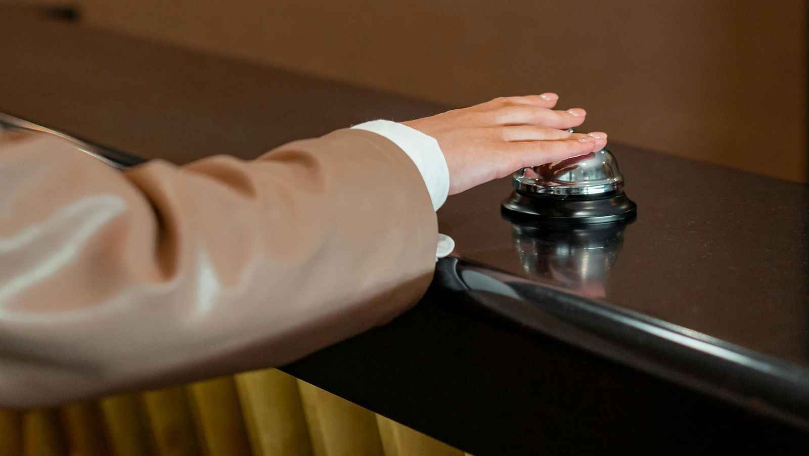A hand in a light brown sleeve presses down on a silver reception bell on a dark counter.