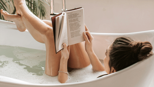 Woman relaxing in a bathtub with bubble bath, reading a book near a large window with natural light.