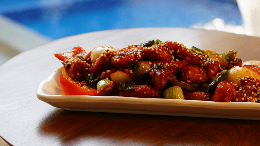A single plate of prepared food sits on a table with a blurred view of a swimming pool at Bougainvillea, Alibaug.
