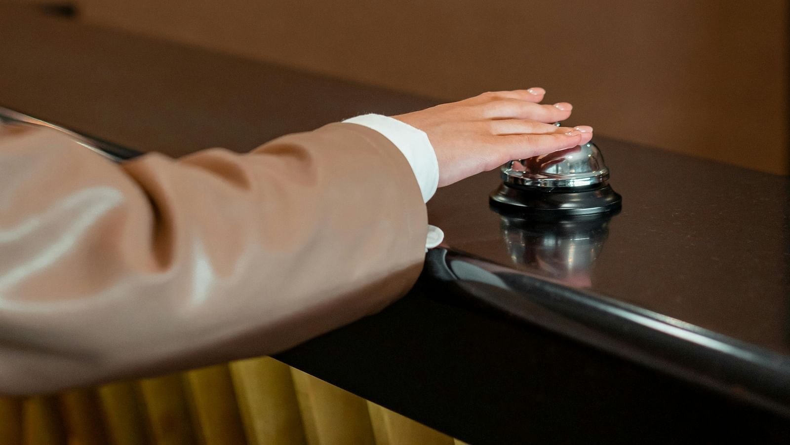 A hand in a light brown sleeve presses down on a silver reception bell on a dark counter.