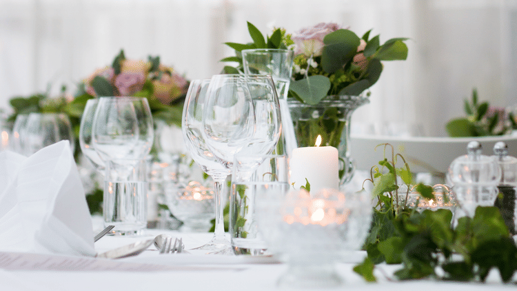 Elegant wedding table setup with glassware, floral centerpieces, greenery, and candles under a white canopy with a hanging chandelier above