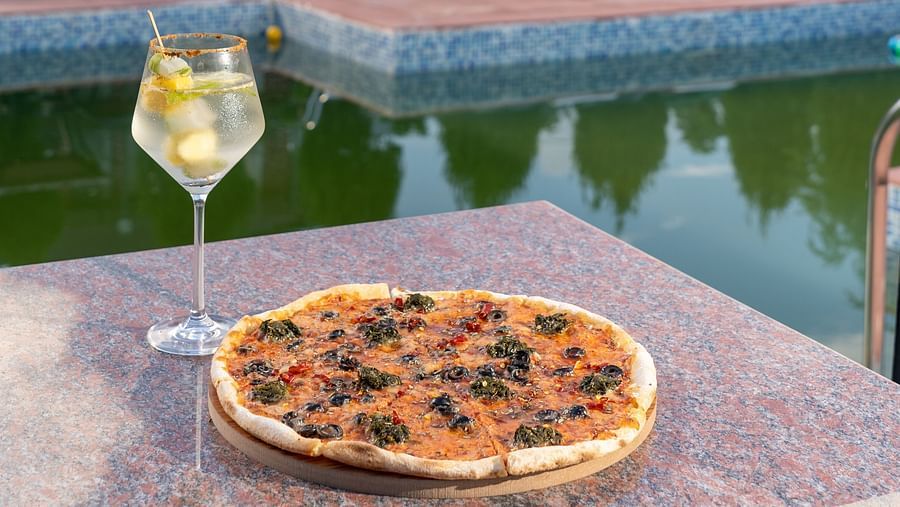 pizza served with a cold beverage with views of the swimming pool and resort in the backdrop - Neer Ganga Resorts