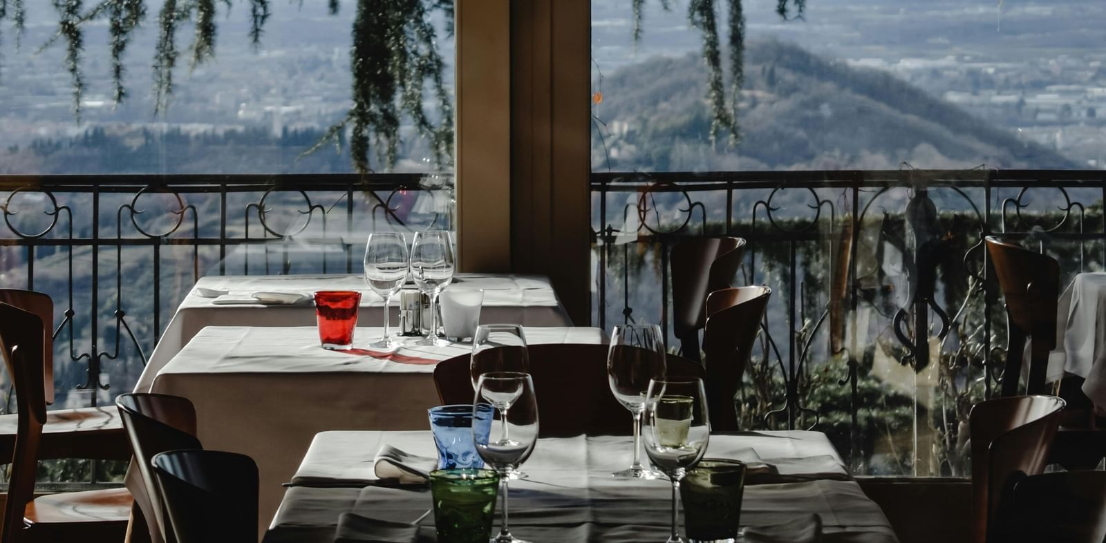 Indoor restaurant dining area with set tables, glassware, and a scenic view of mountains and trees visible through large windows in the background.