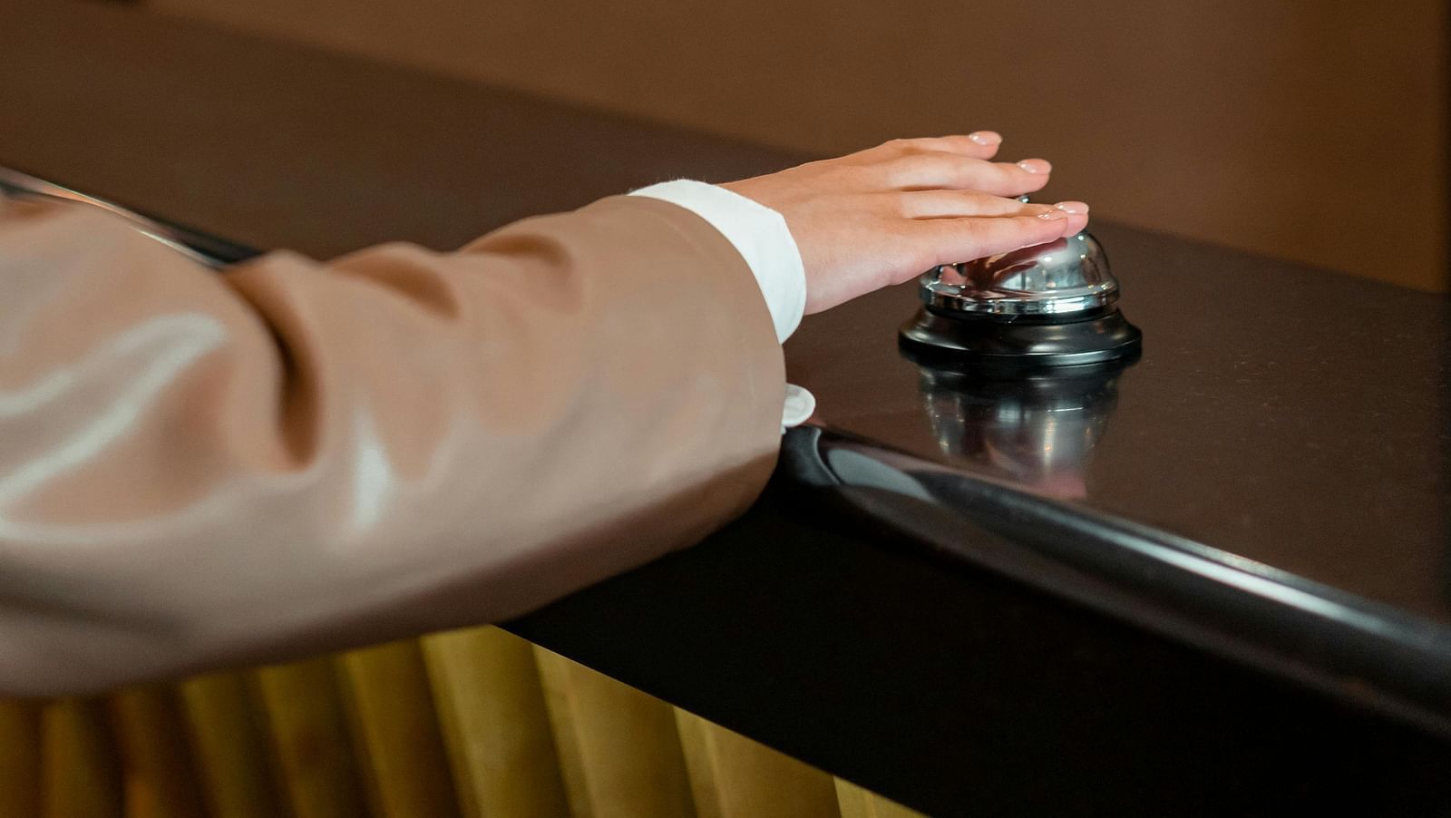 A hand in a light brown sleeve presses down on a silver reception bell on a dark counter.