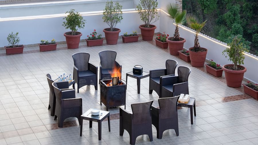 A rooftop terrace at Clarks Inn, Near Kainchi Dham features a fire pit with seating, potted plants, and string lights, overlooking hills, trees, and some buildings.