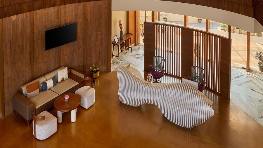 An elevated view captures a hotel lobby with a striking white, undulating reception desk. Above, a modern chandelier hangs from a ceiling with exposed wooden beams, while seating areas are arranged on the polished floor below - Voyage The Solitaire Bliss.