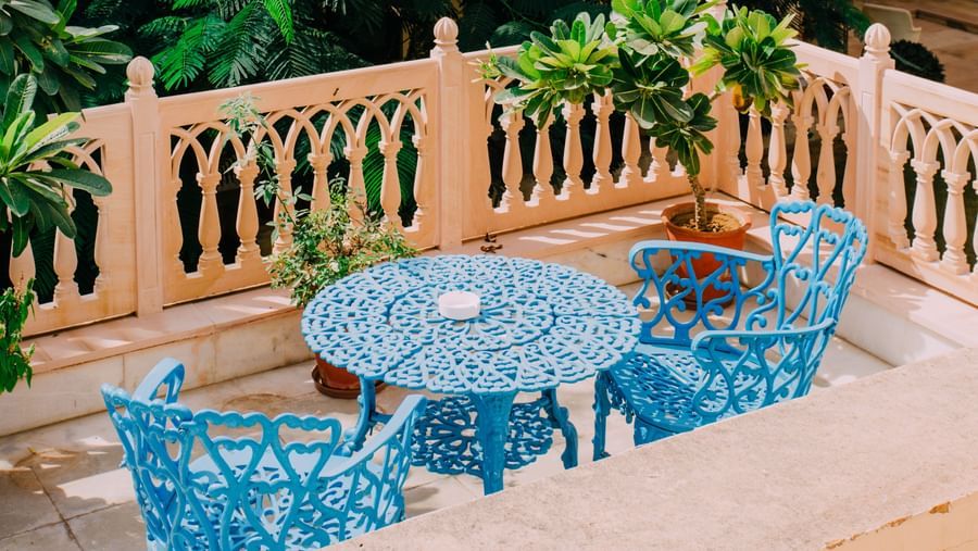 A balcony or terrace at Royal Heritage Haveli with a blue table and chairs, enclosed by a railing and surrounded by dense green foliage.