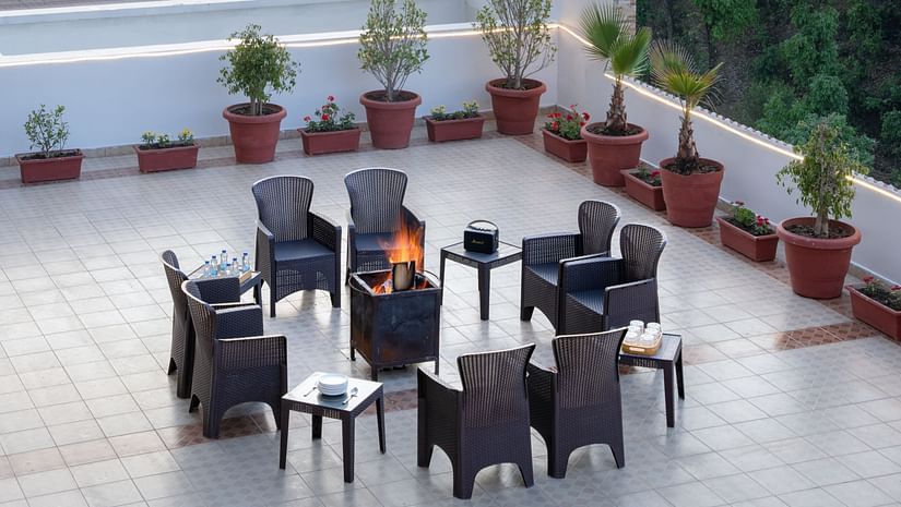 A rooftop terrace at Clarks Inn, Near Kainchi Dham features a fire pit with seating, potted plants, and string lights, overlooking hills, trees, and some buildings.