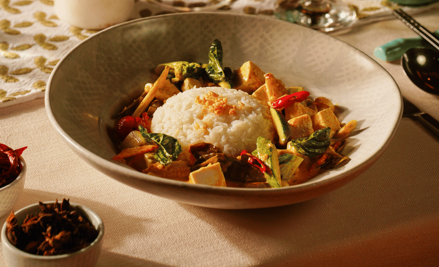 A bowl of curry with rice, tofu, and vegetables on a table with a glass of wine, water, a candle, and bamboo steamer. Warm, elegant dining scene.