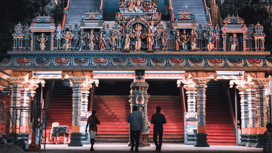 A night view of a brightly lit, multi-tiered temple structure with people visible at the base.