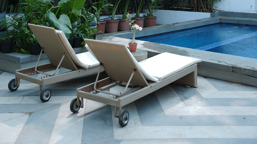 2 wooden sun loungers placed on a tiled deck next to a swimming pool and thick green foliage at Bougainvillea, Alibaug.