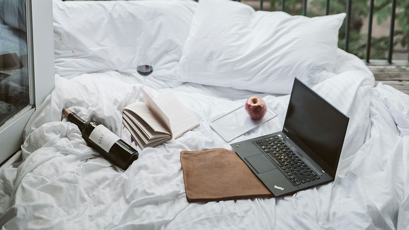 A bed setup on a balcony with white pillows and blanket, featuring a laptop, book, wine bottle, glass of wine, apple, and a peaceful green view. 