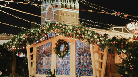 A beautifully decorated Christmas market glows with fairy lights beneath a tall, illuminated tower under the night sky.