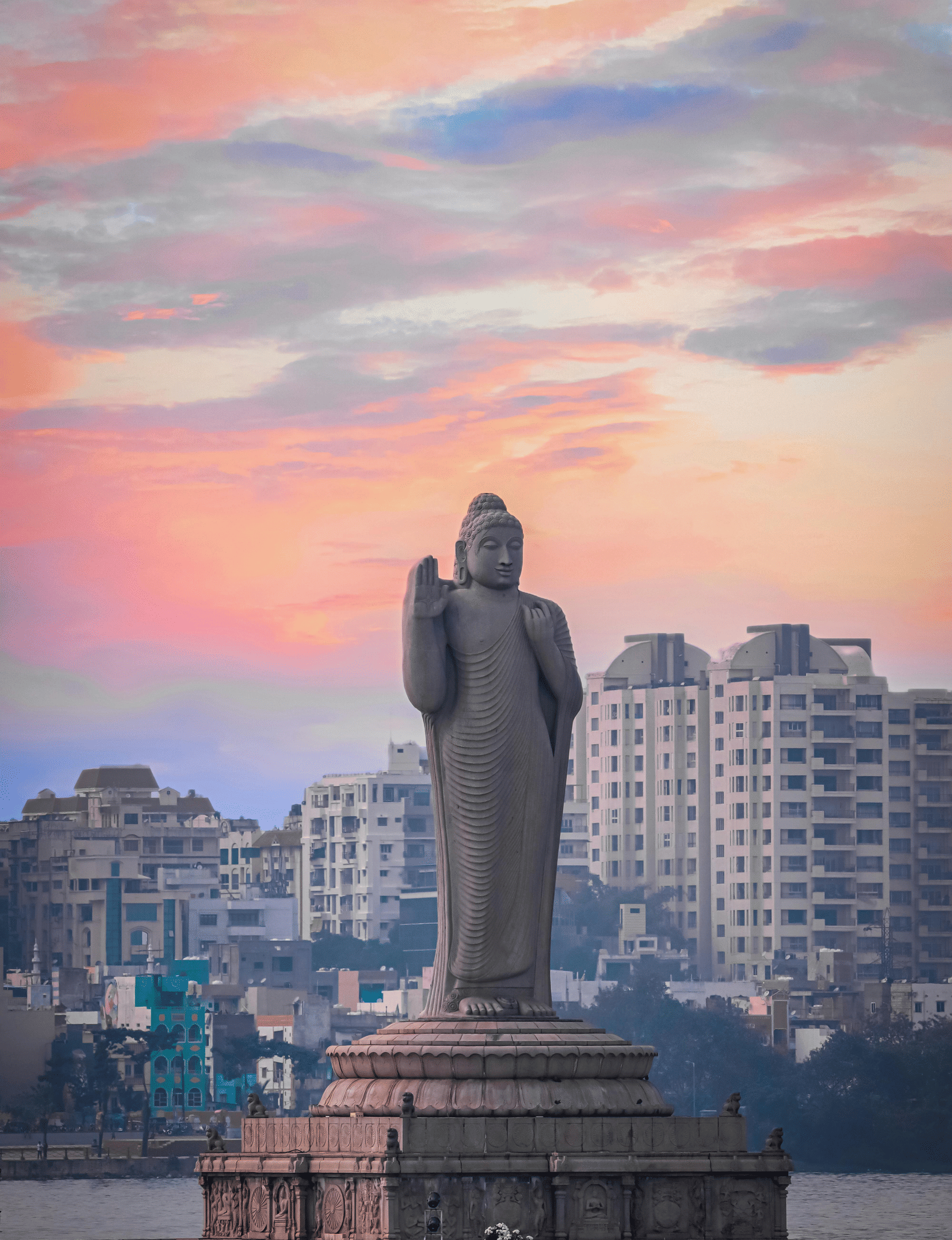 A Budha Statue made of stone located in the middle of a water body under a sky with orange and pink hues.