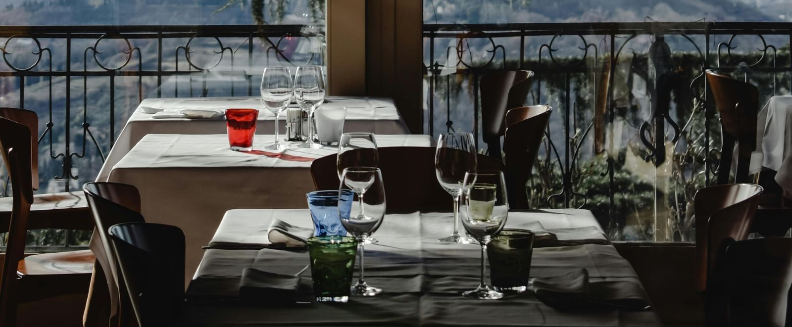 Indoor restaurant dining area with set tables, glassware, and a scenic view of mountains and trees visible through large windows in the background.