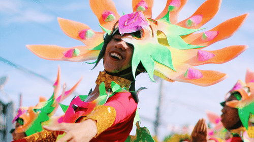 Street festival performers in colourful costumes with orange and green feather-shaped headdresses, red and gold outfits, dancing in a parade.