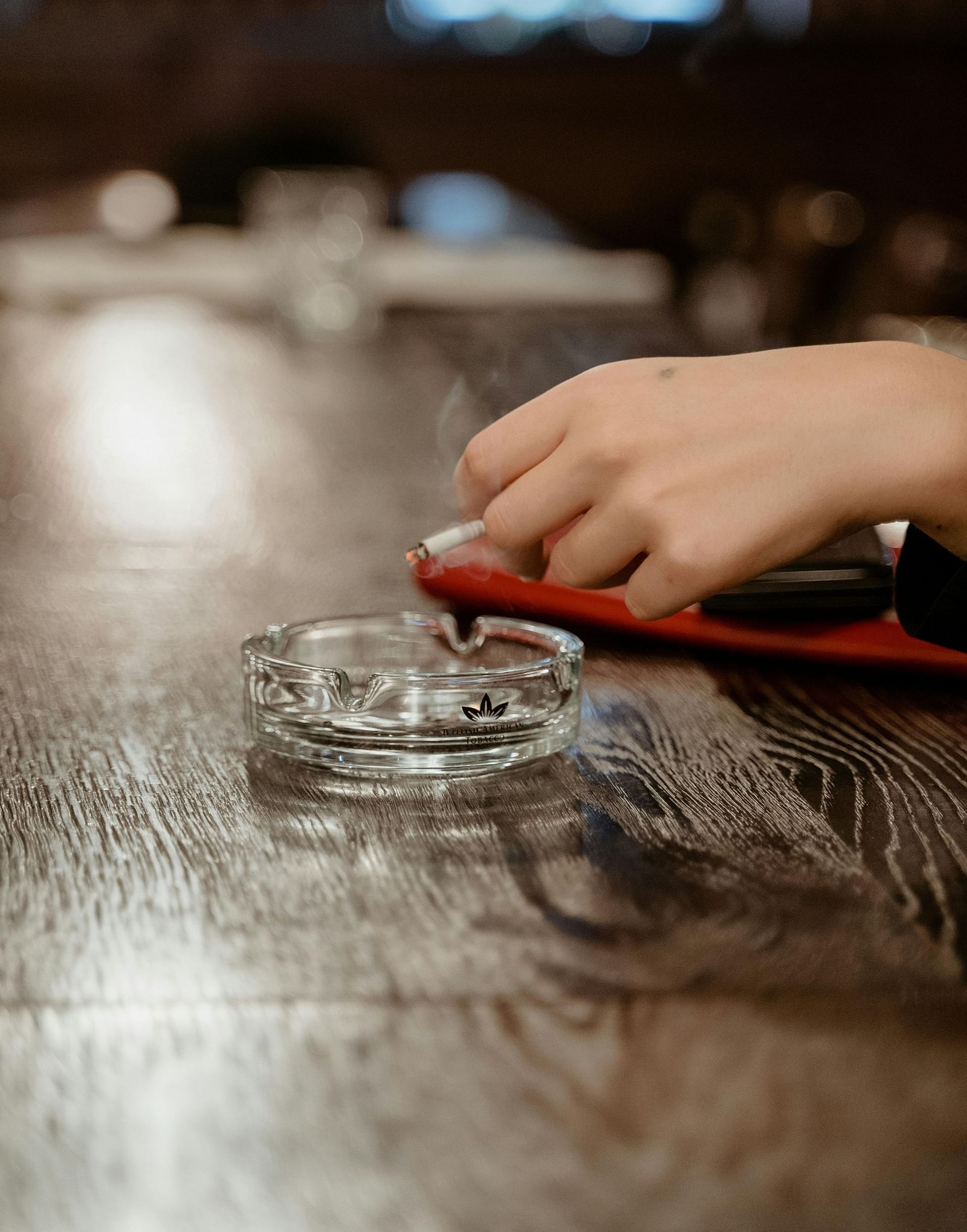 A person holding a cigarette over a crystal ash tray kept on a counter. 