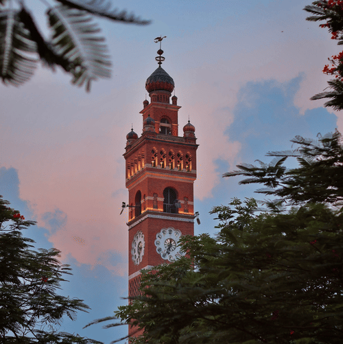The illuminated facade of the Hussainabad Clock Tower, Lucknow, during the late evening hours with the canopy of trees in the foreground covering the tower in view.