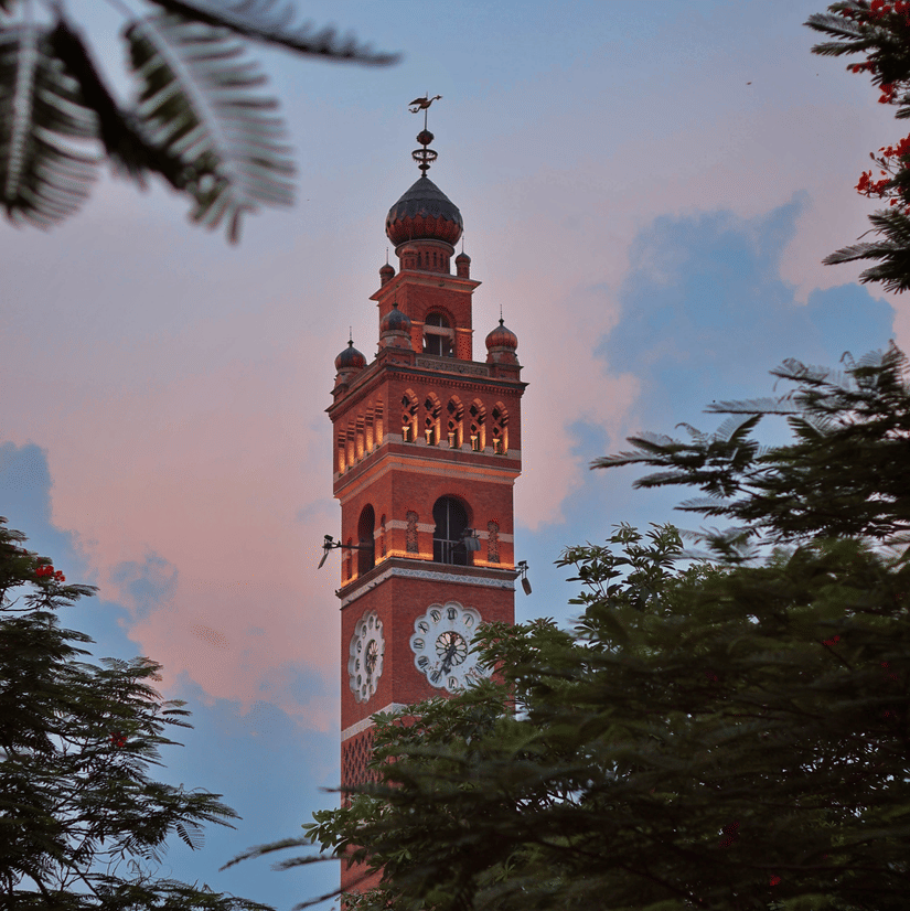 The illuminated facade of the Hussainabad Clock Tower, Lucknow, during the late evening hours with the canopy of trees in the foreground covering the tower in view.