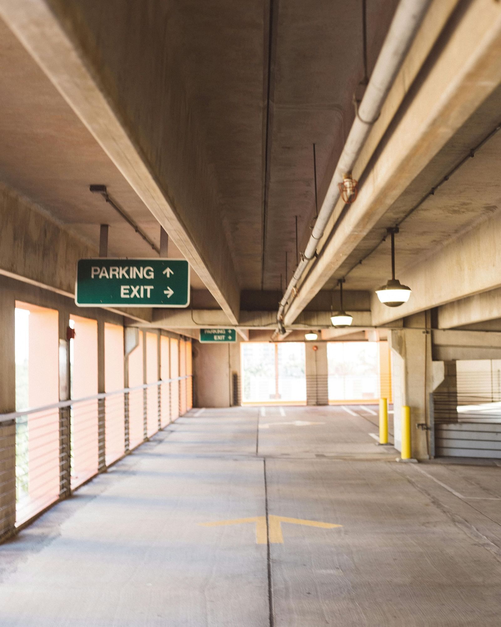 A view looking down a covered walkway with a concrete floor and ceiling, showing a green sign that reads lobby and exit with arrows pointing in different directions, and natural light visible at the far end.