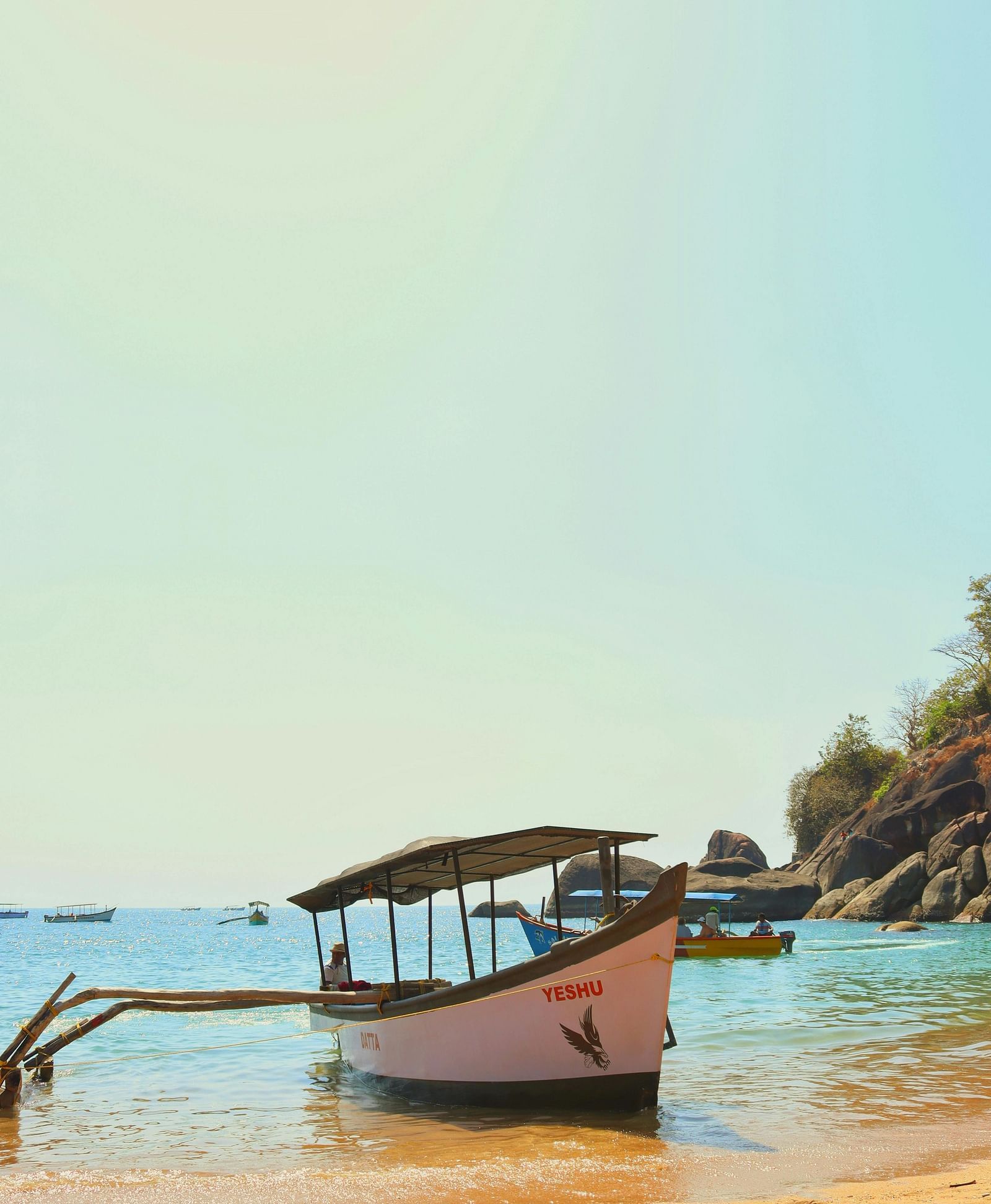 A boat named Yeshu beached on a sandy shore with clear waters and rocky outcrops in the background-15610234
