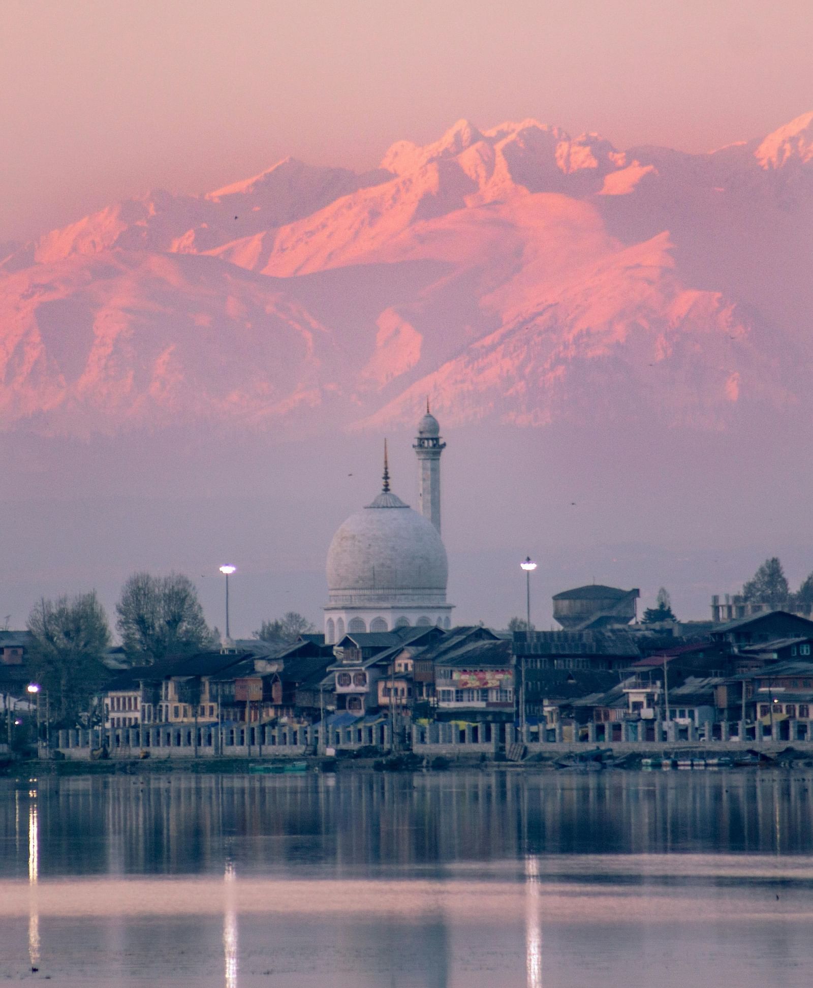 A pink-hued early morning view of the snow-capped Himalayas overlooking a calm lake with a white dome and minaret in the foreground