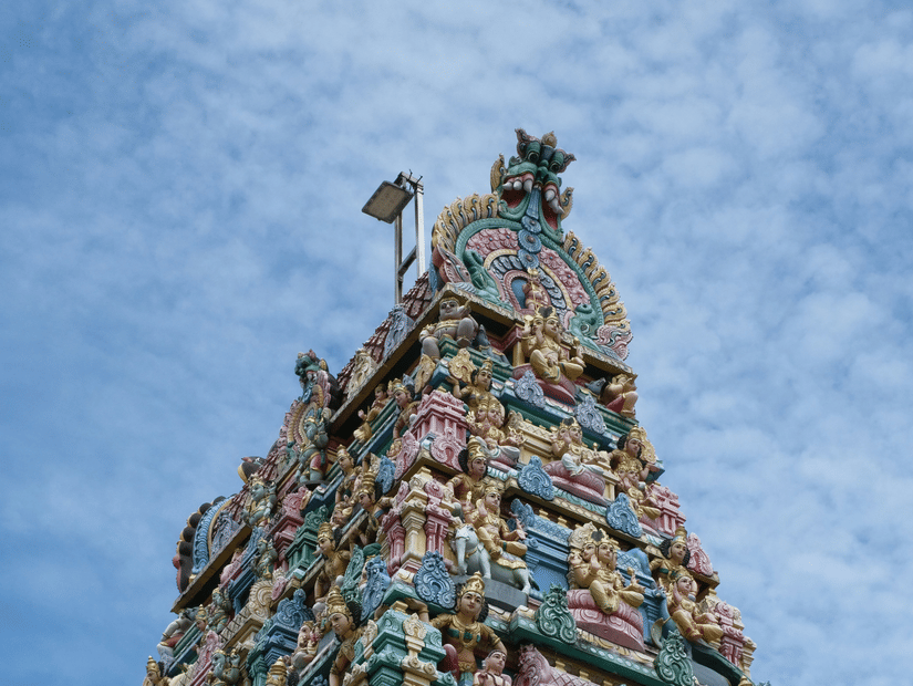 A view from below of Pillayarpatti Ganapathi temple's facade with numerous intricate small deities carved into the gopuram and blue sky in the background..