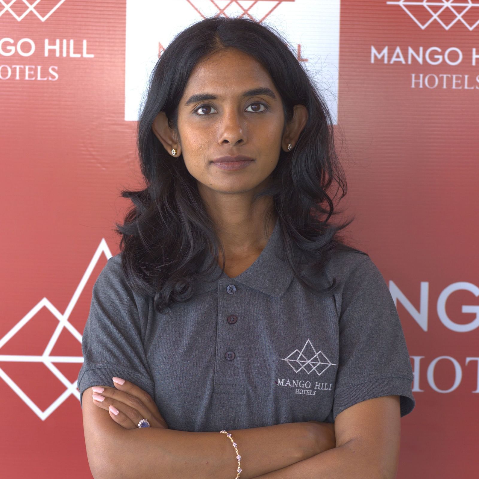 Gayathiri Om in a grey Mango Hill Hotels uniform standing with arms crossed in front of a branded backdrop.