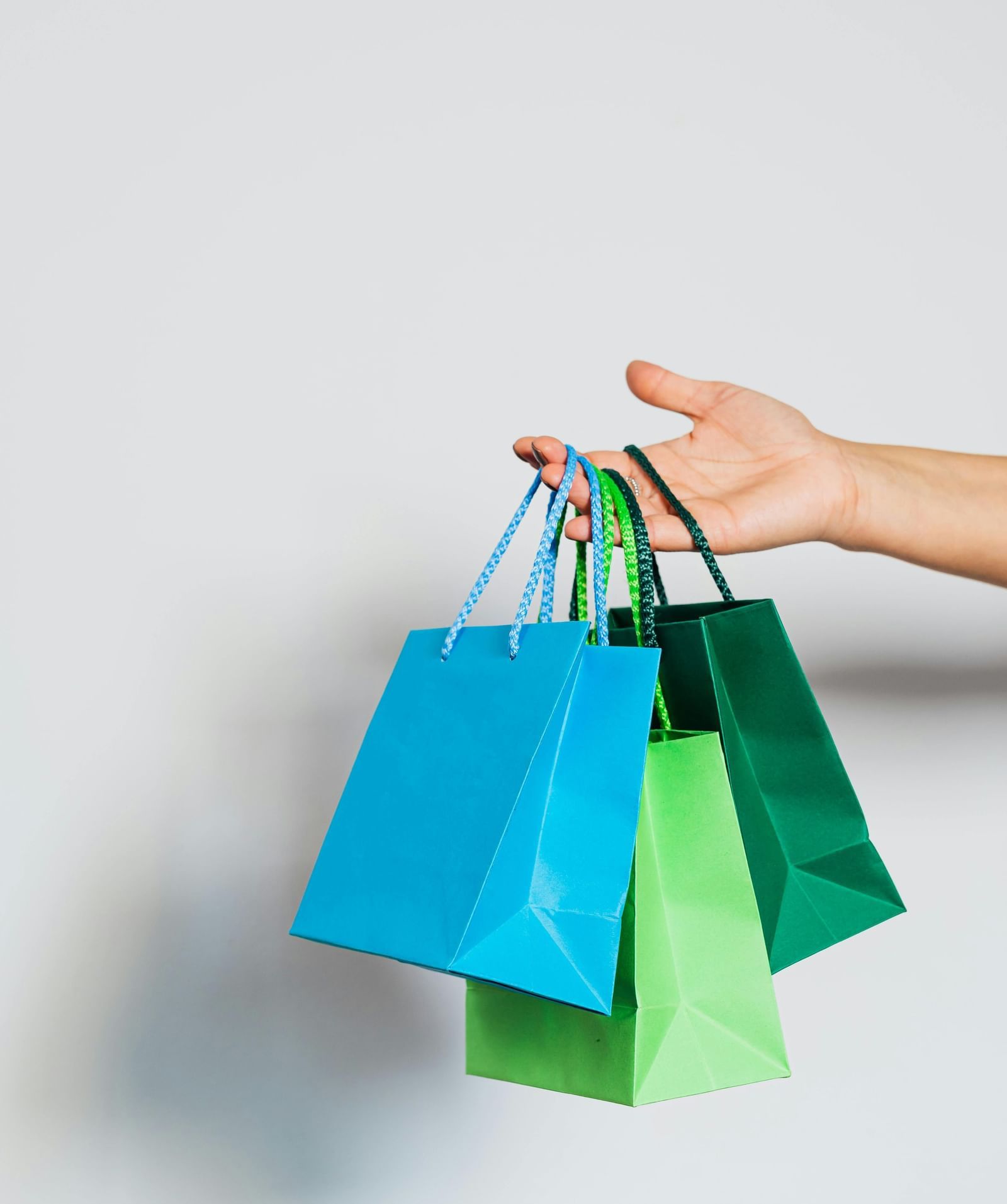 A person's hand stretched out, holding three paper shopping bags with a plain wall in the background.