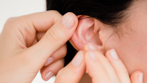 2 hands are shown adjusting or placing something onto a person's ear, with a white background.