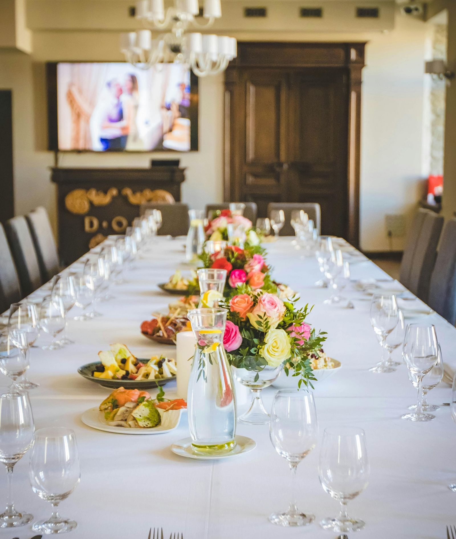 A long dining table decorated with flowers in the middle along with shiny glassware and cutlery set on the table.