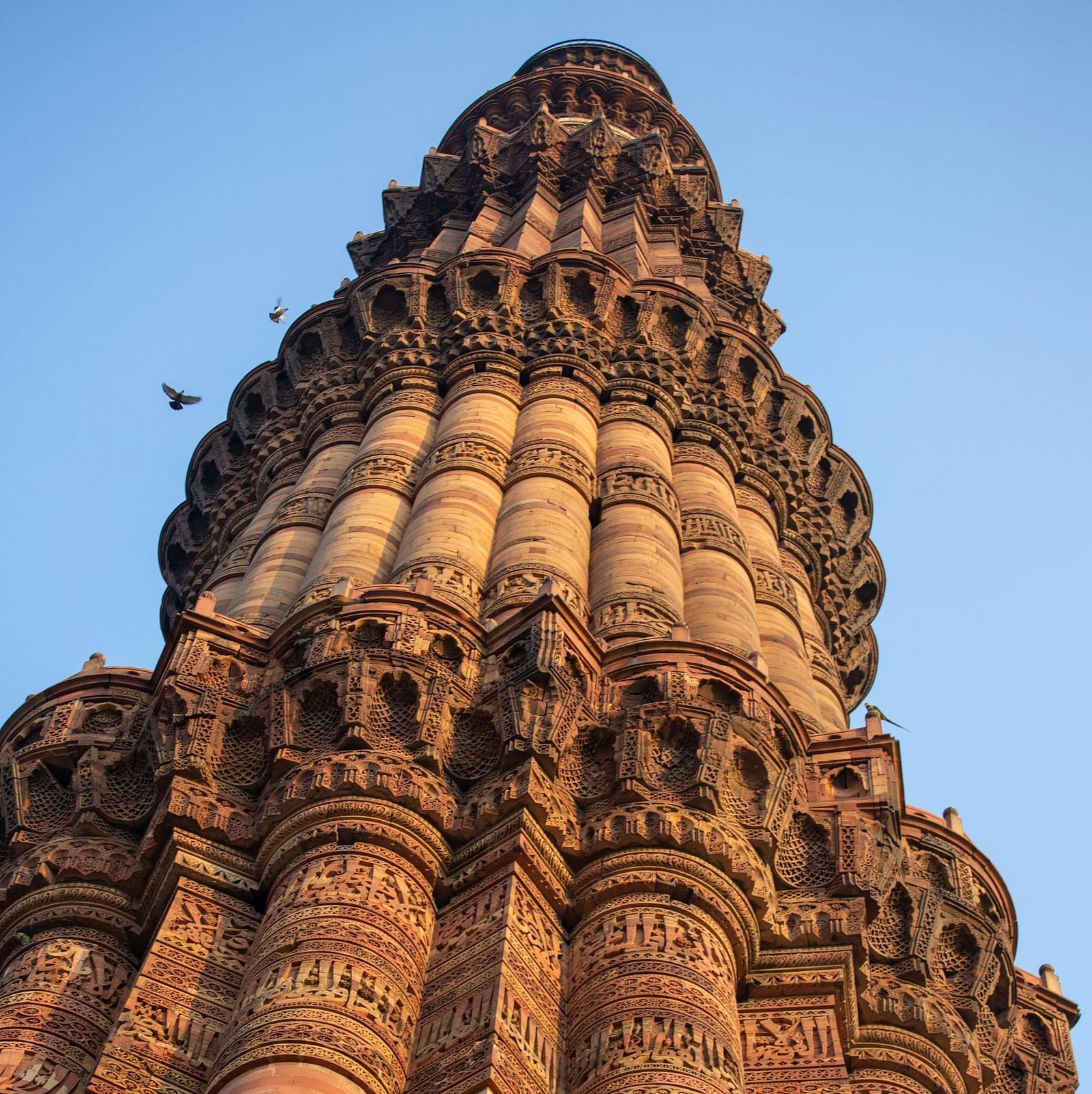 A low-angle view of the Qutub Minar, a tall, intricately carved, reddish-brown minaret, stands against a clear blue sky.