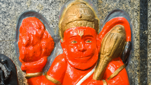 A close up of the Hindu deity Hanuman in orange and gold colour, and a stone in the background.