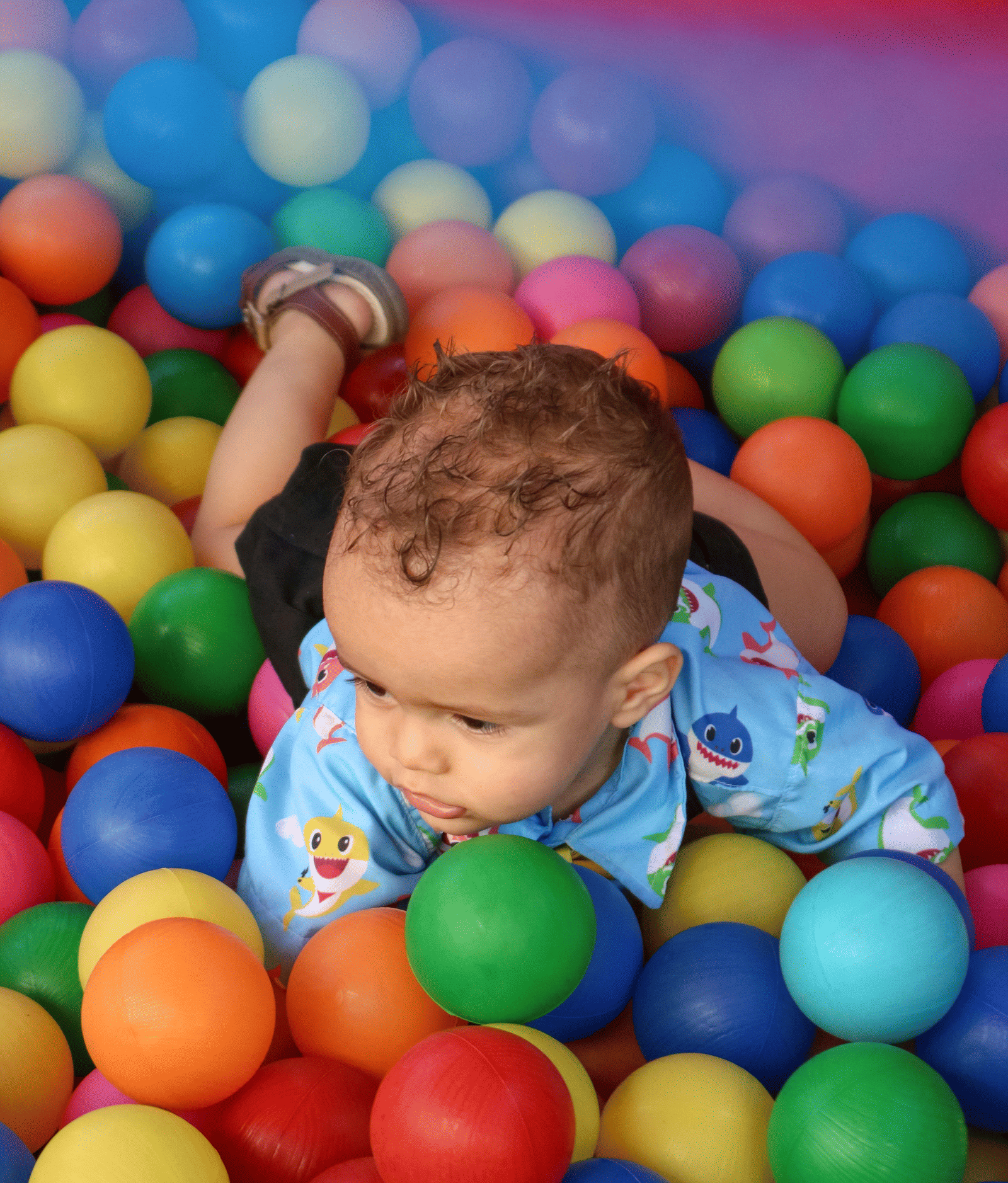 A young baby in a blue onesie smiles while lying in a bright ball pit filled with multi-colored plastic balls.