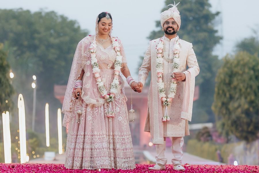 Bride and groom holding hands while walking on a pink flower-decorated platform over water.