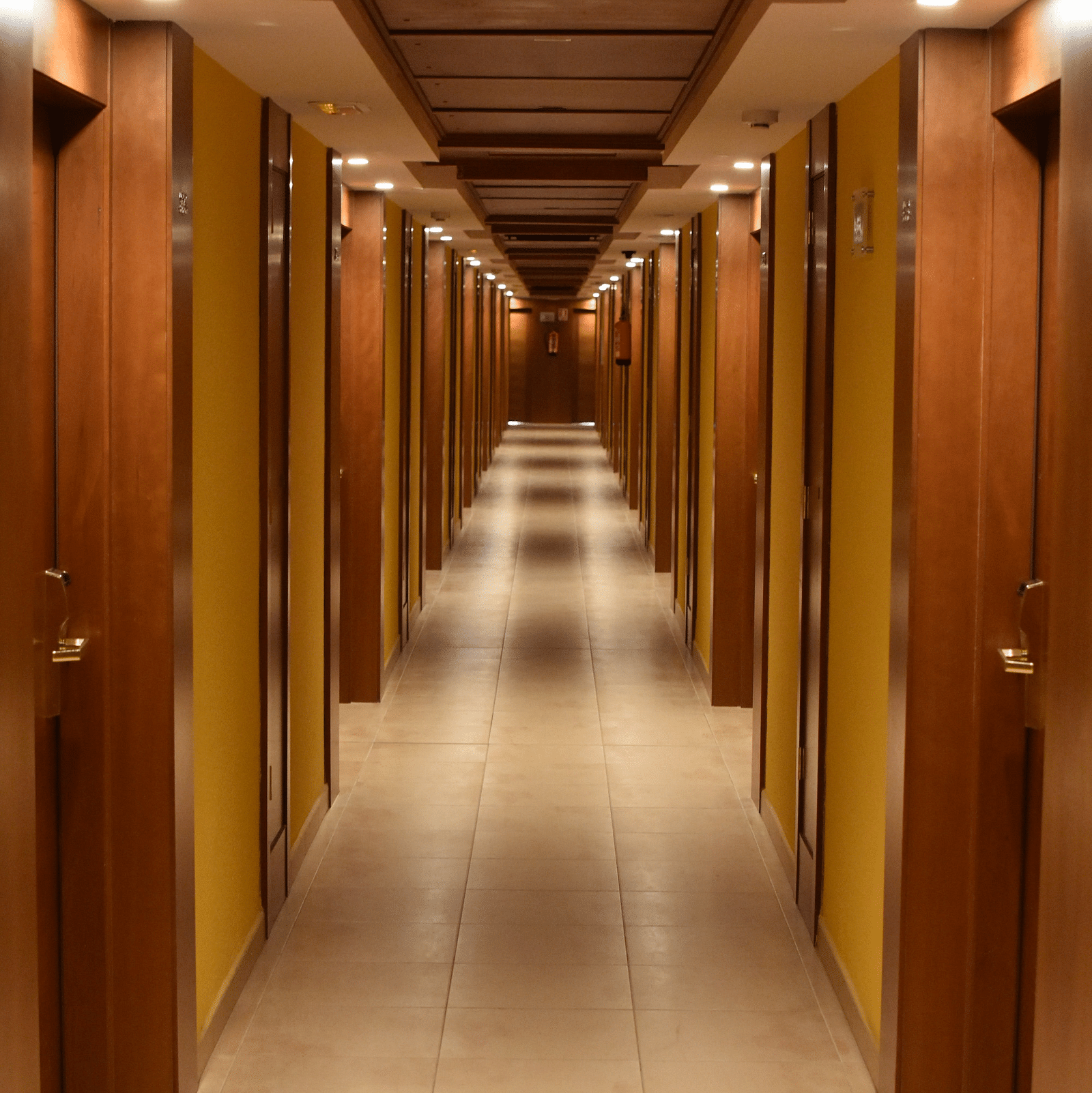 A narrow hotel corridor lined with doors and yellow walls, illuminated by recessed ceiling lights
