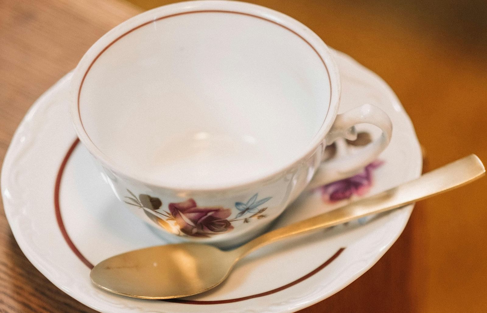 A white porcelain teacup with floral patterns placed on a matching saucer, accompanied by a gold spoon, resting on the edge of a wooden surface.