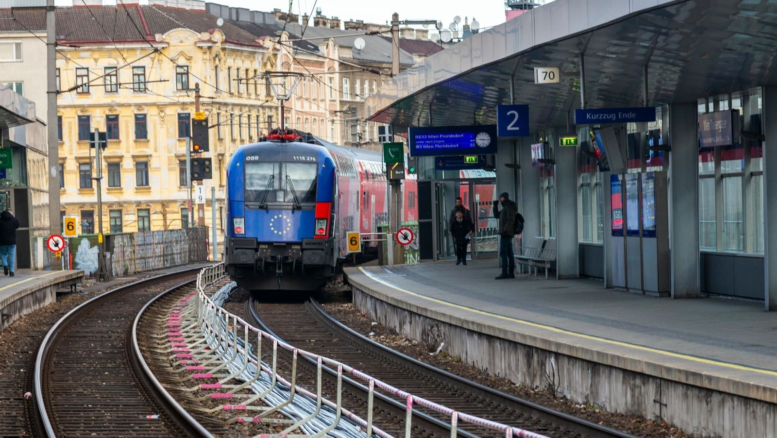 A low-angle view of a train station with a train approaching on a curved track. A person stands on the platform under an overhang on the right.