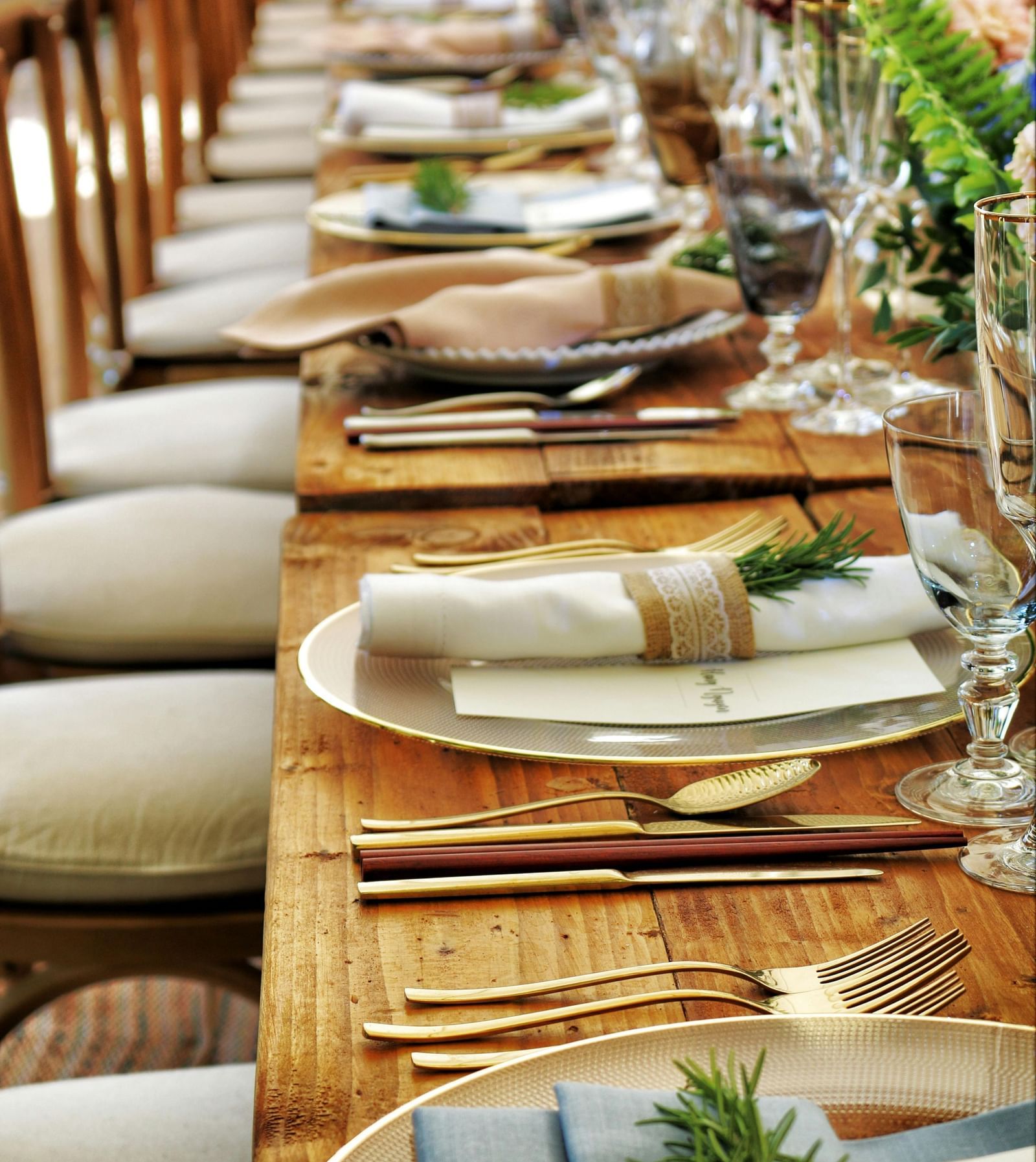 A long, wooden table is set for a rustic wedding reception, featuring neutral-toned place settings with natural accents like rosemary sprigs and twine-wrapped napkins, complemented by elegant glassware and floral arrangements. 