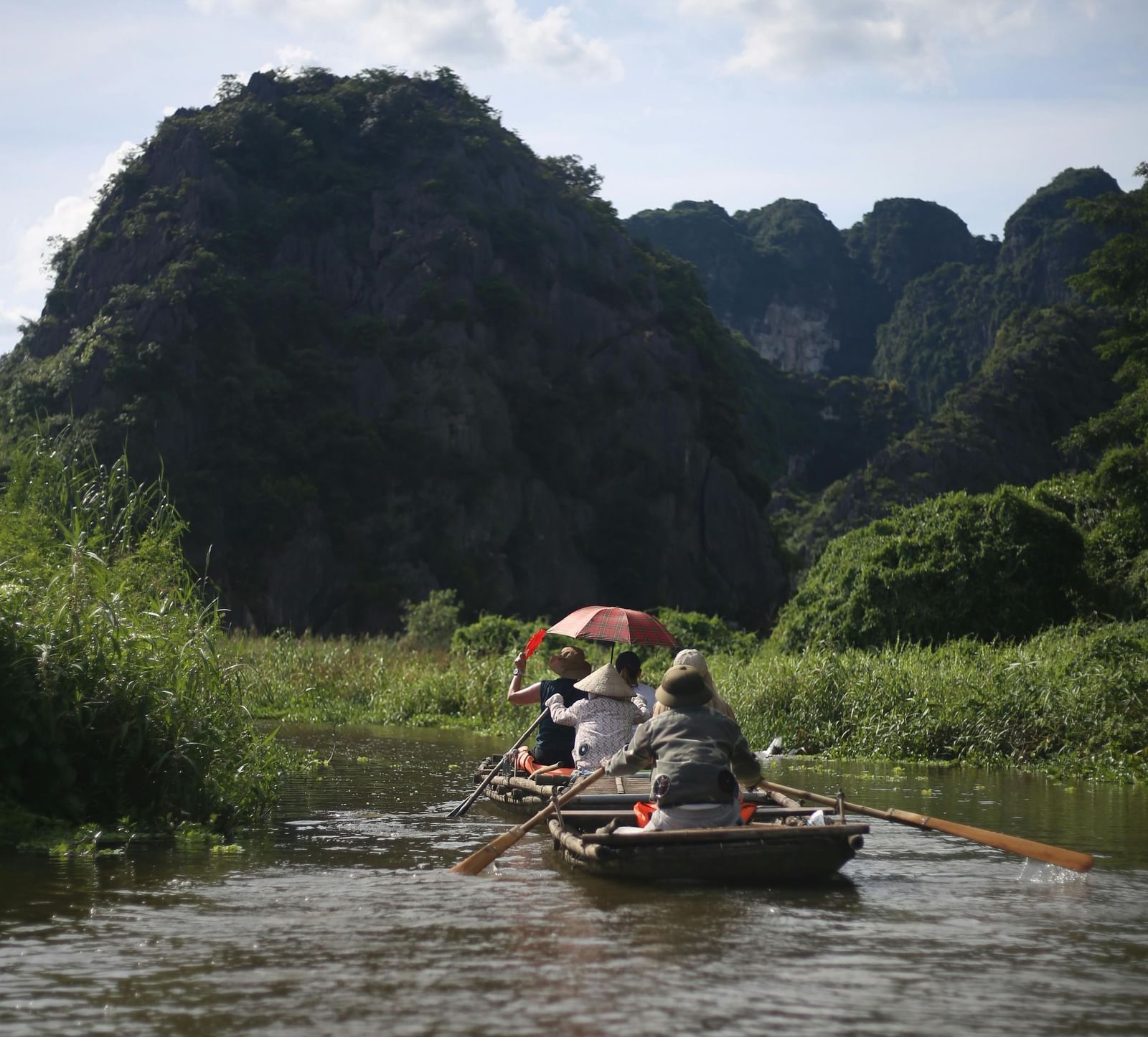 pexels-People rowing a wooden boat along a river surrounded by greenery, with tall rocky hills in the background under a partly cloudy sky.robin-stolk-1052962922-33505245
