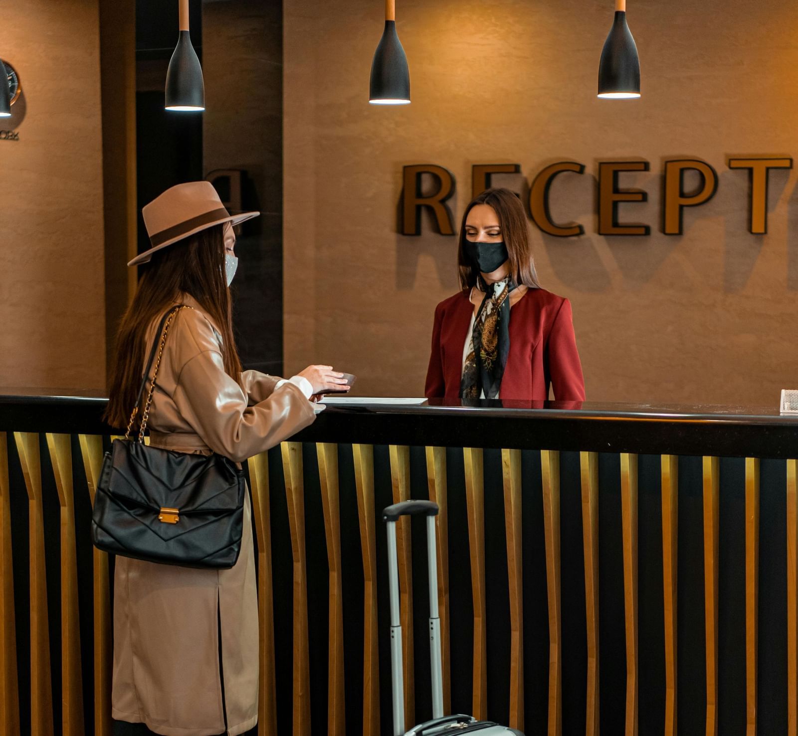 A receptionist standing behind the desk greeting the guest carrying a luggage trolley in front of her.