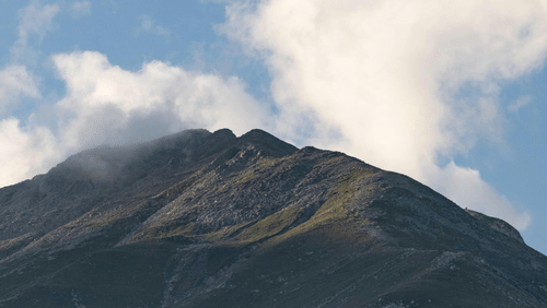 A majestic mountain peak under a partly cloudy sky, with sunlight illuminating a patch of its grassy slope, contrasting with the darker forested base.